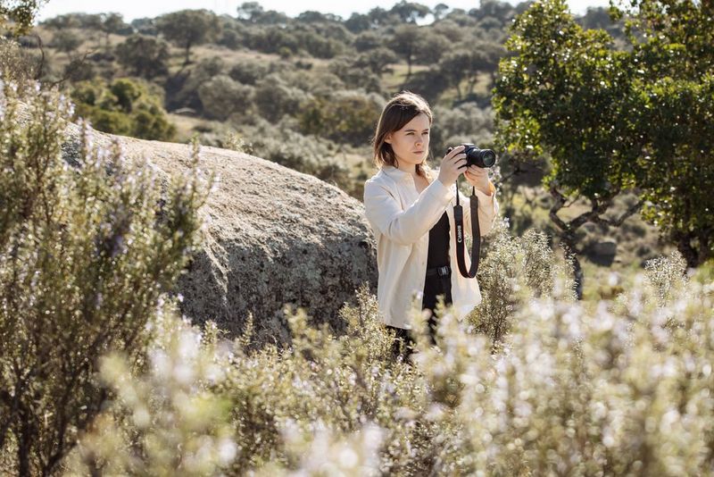 A woman using a Canon EOS R7, with trees and bushes around her and more visible in the distance.