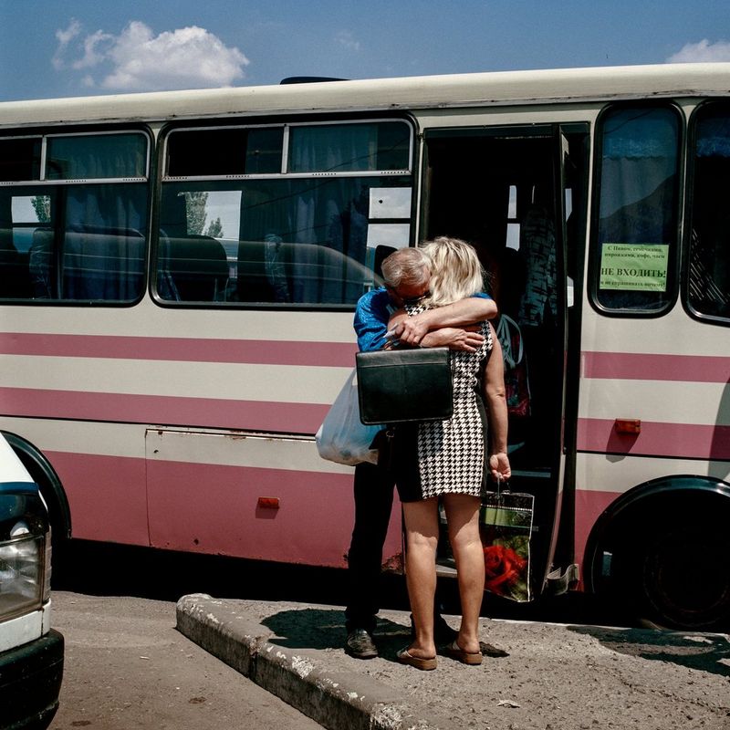  An old white man embraces his wife beside a red and white bus in Donbas, Ukraine. Image taken by Anastasia Taylor-Lind.