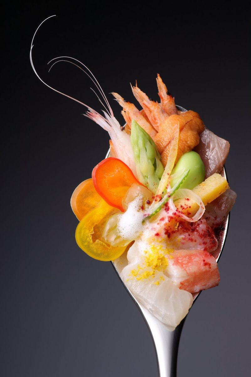 Ряд небольших ингредиентов, включая побег спаржи и хвост креветки, разложены на ложке.