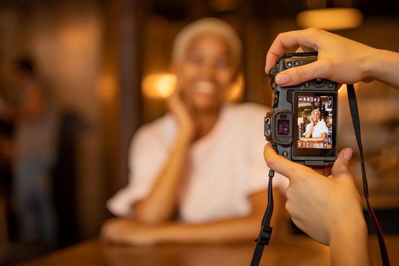  A photographer&#39;s hands hold a Canon EOS RP as she photographs a smiling woman in a dimly-lit interior.