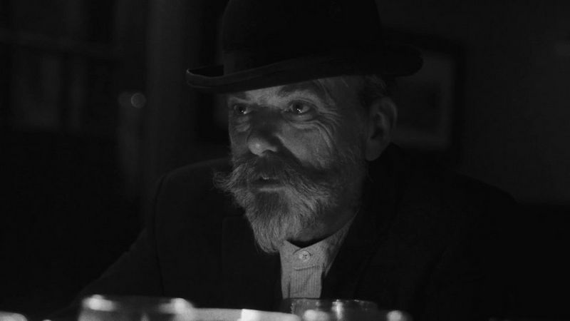 Close up black and white shot of one of the actors, an older man with a mustache and beard wearing an old fashioned hat