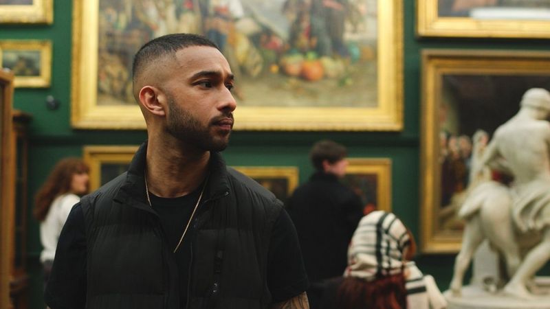 Young man stood in an art gallery in front of some paintings on the wall, looking to the side 