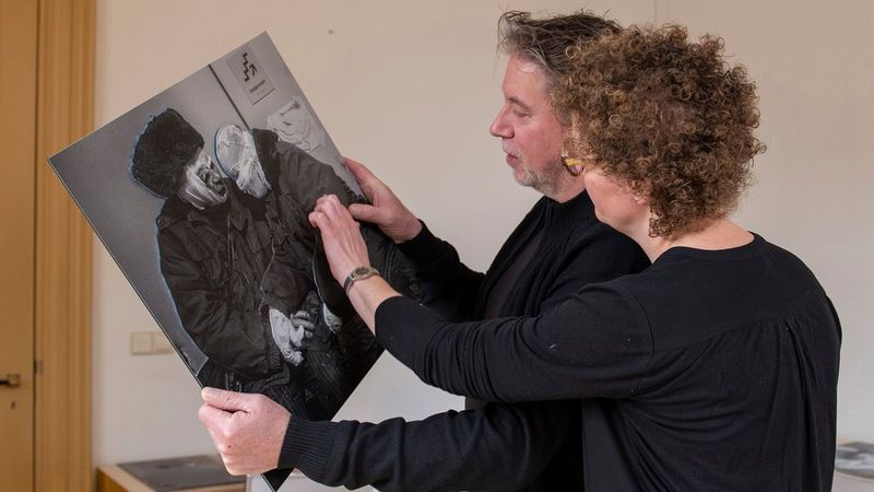 A man and a woman stand side by side, inspecting a large black and white photograph of two people, huddled together.