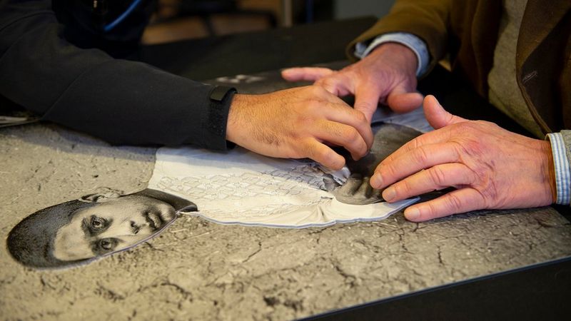 Two pairs of hands explore a black and white elevated print image of a young man.