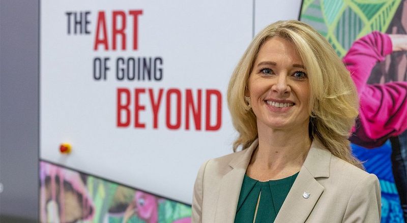 Jennifer Kolloczek is smiling and standing in front of a sign which reads ‘The Art of Going Beyond’