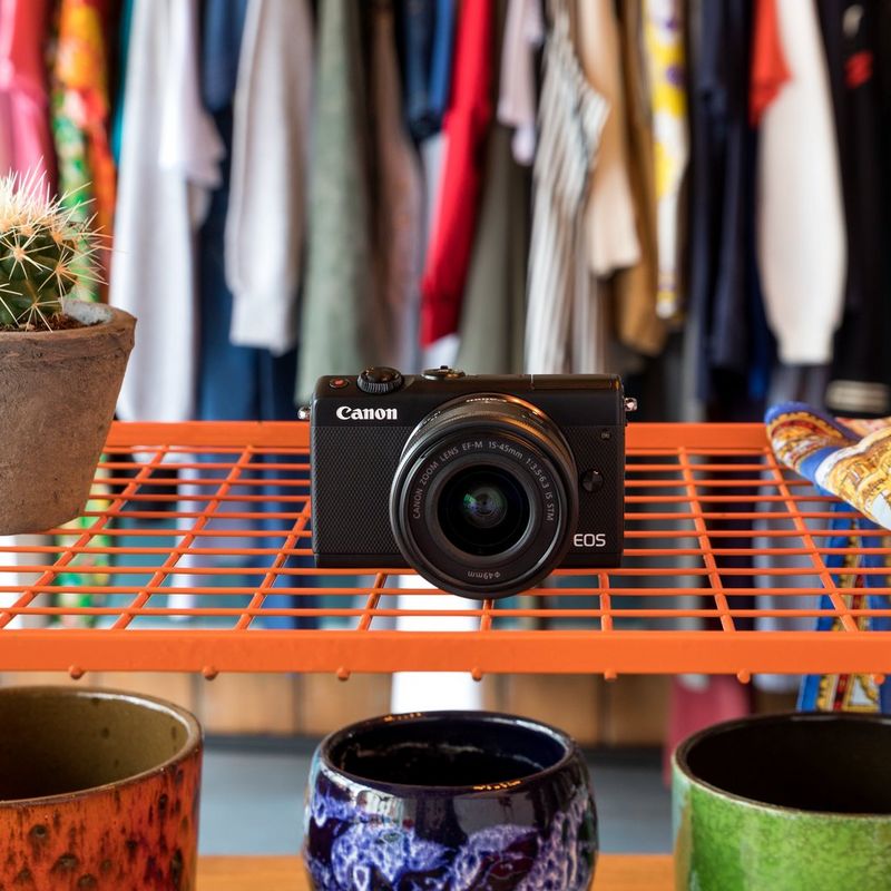 Canon EOS M100 Schwarz + Canon EF-M 15-45mm f/3.5-6.3 IS STM Objektiv Schwarz Produkt Diagonale Ansicht mit internem Blitz und klappbarem Display von hinten