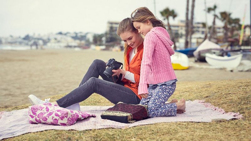 A mother and daughter on a sandy beach look at the screen of a Canon EOS 250D.