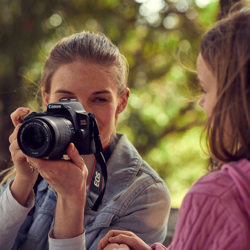 Canon EOS 250D Gehäuse, Silber + EF-S 18-55mm f/4-5.6 IS STM Objektiv Frau, die ein Foto aufnimmt, mit Vorderansicht des Produkts