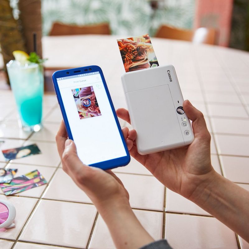 Canon Zoemini S Sofortbildkamera und Mini-Fotodrucker, Perlweiß Drucker, aus dem ein Druck kommt, mit einer Kamera in der anderen Hand