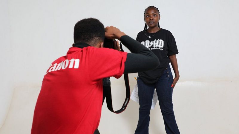 A photographer in a red Canon shirt aims a Canon camera at a young woman who is posing in a black t-shirt and jeans against a white background.