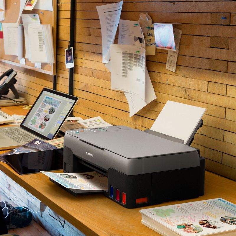 Canon PIXMA G2520 3-in-1 MegaTank Farbdrucker mit nachfüllbaren Tintenbehältern Auf einem Bürotisch neben einem Laptop und mit ausgeklapptem Papierfach und Papierstütze