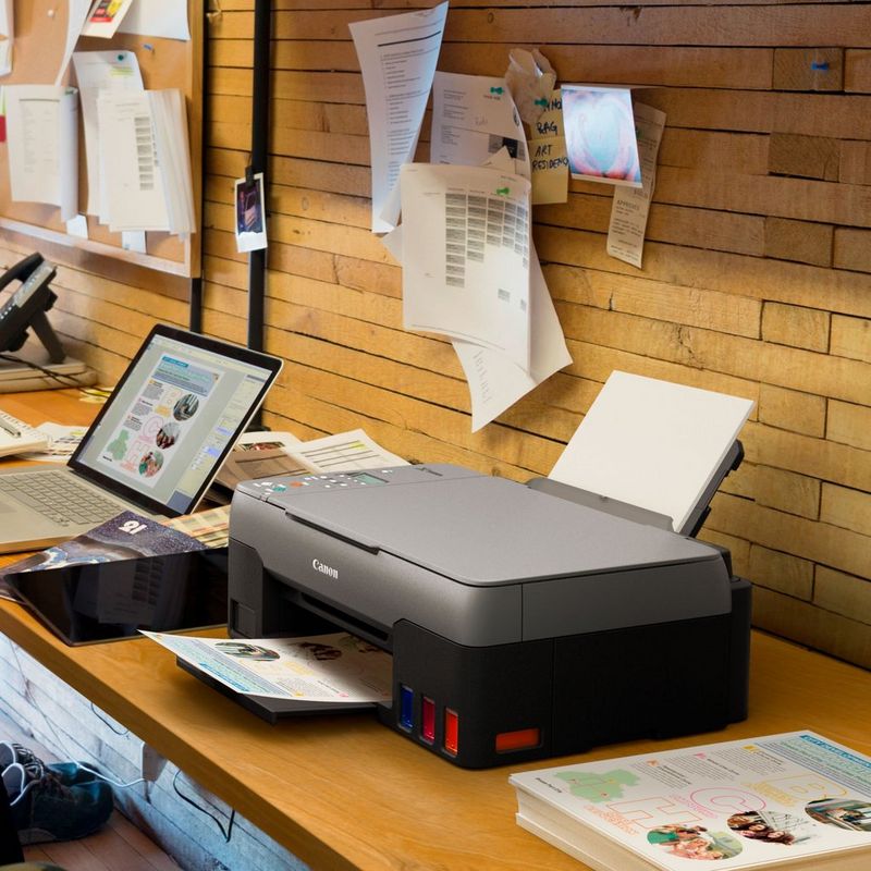Canon PIXMA G3520 3-in-1 drahtloser MegaTank Farbdrucker mit nachfüllbaren Tintenbehältern Auf einem Bürotisch neben einem Laptop und mit ausgeklapptem Papierfach und Papierstütze