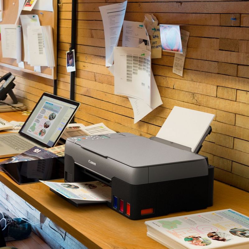 Canon PIXMA G3560 3-in-1 drahtloser MegaTank Farbdrucker mit nachfüllbaren Tintenbehältern Auf einem Bürotisch neben einem Laptop und mit ausgeklapptem Papierfach und Papierstütze