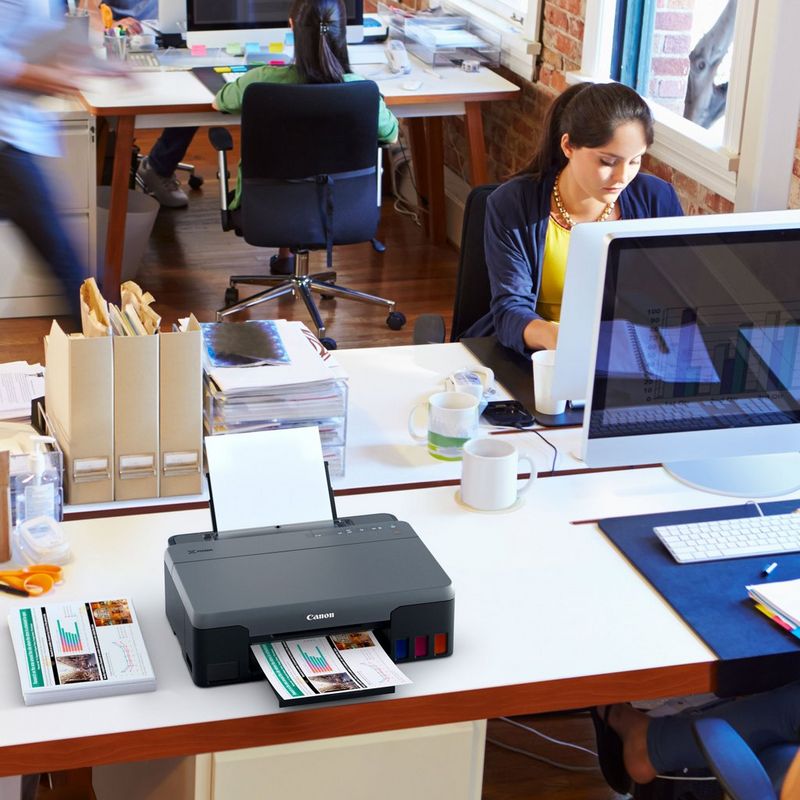 Canon PIXMA G1520 MegaTank Farbdrucker mit nachfüllbaren Tintenbehältern Auf einem Bürotisch neben einem Desktop-PC und mit ausgeklapptem Papierfach und Papierstütze