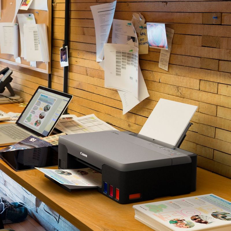 Canon PIXMA G1520 MegaTank Farbdrucker mit nachfüllbaren Tintenbehältern Auf einem Bürotisch neben einem Laptop und mit ausgeklapptem Papierfach und Papierstütze