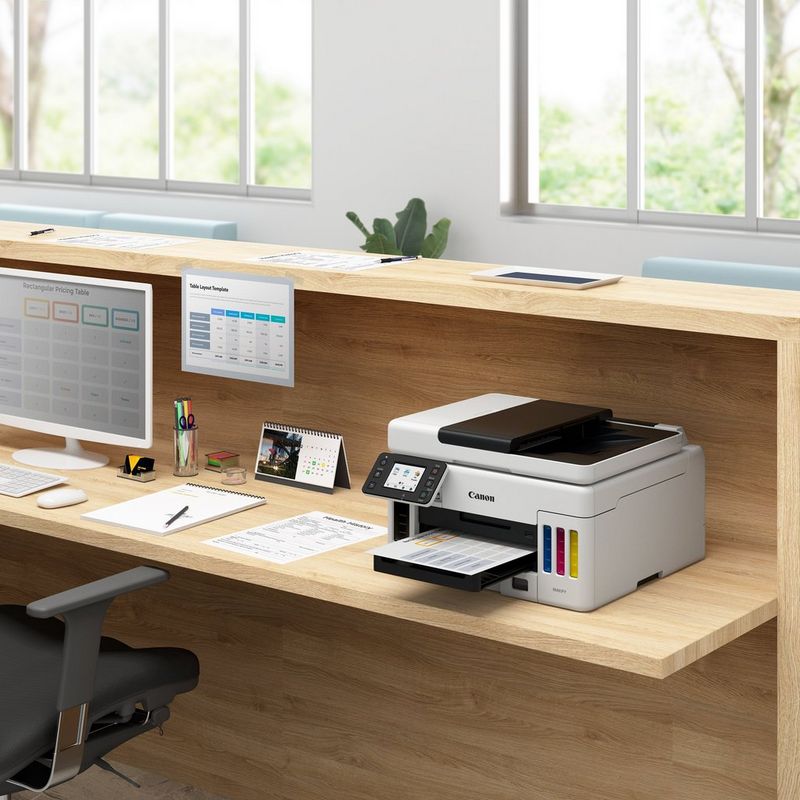 MAXIFY GX6050 MegaTank 3-in-1 Tintenstrahl-Multifunktionssystem mit nachfüllbaren Tintentanks und WLAN Auf einem Bürotisch neben einem Desktop-PC und mit ausgeklapptem Papierfach