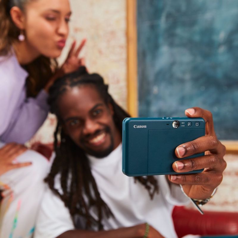 Canon Zoemini S2 Sofortbildkamera mit Mini-Fotodrucker, Aquamarin + Tagebuch + Tasche Paar knipst ein Selfie mit einer Tafel im Hintergrund