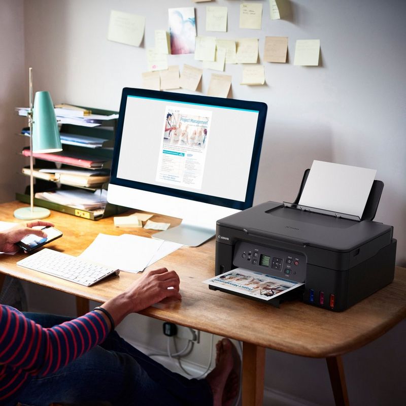 Canon PIXMA G3570 WLAN 3-in-1 MegaTank Farb-Multifunktionssystem mit nachfüllbaren Tintentanks , Schwarz Produkt auf einem Schreibtisch, an dem eine Frau ein Telefon in der Hand hält