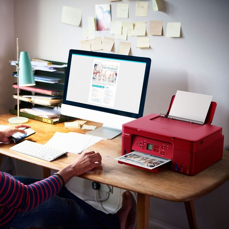 Canon PIXMA G3572 WLAN 3-in-1-Farb-Multifunktionssystem mit nachfüllbarem MegaTank, Rot Produkt auf einem Schreibtisch, an dem eine Frau ein Telefon in der Hand hält