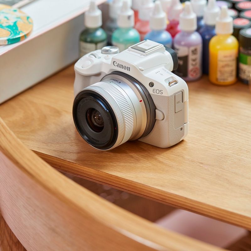 Canon EOS R50 spiegellose Kamera, Weiß und RF-S 18-45mm F4.5-6.3 IS STM Objektiv Produkt auf einem Schreibtisch mit Farbflaschen