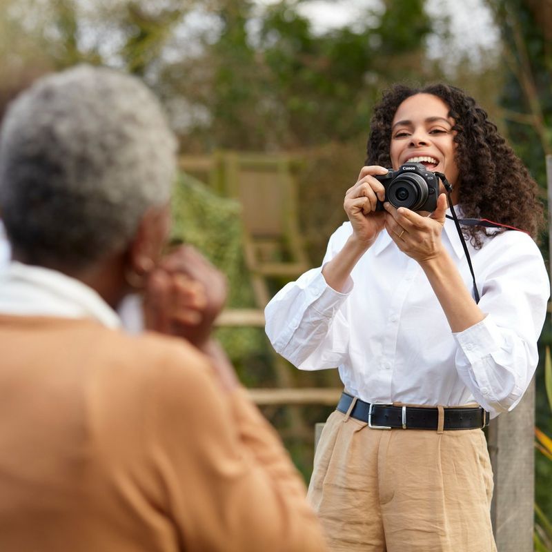 Canon EOS R100 spiegelloses Kamera Frau, die das Produkt mit Tragegurt hält und ein Foto von einem Mann aufnimmt