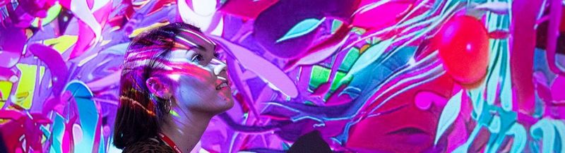 A woman’s head and shoulders as she stands in the middle of a brightly coloured projection of leaves and shapes in pink, red, purple and blue.