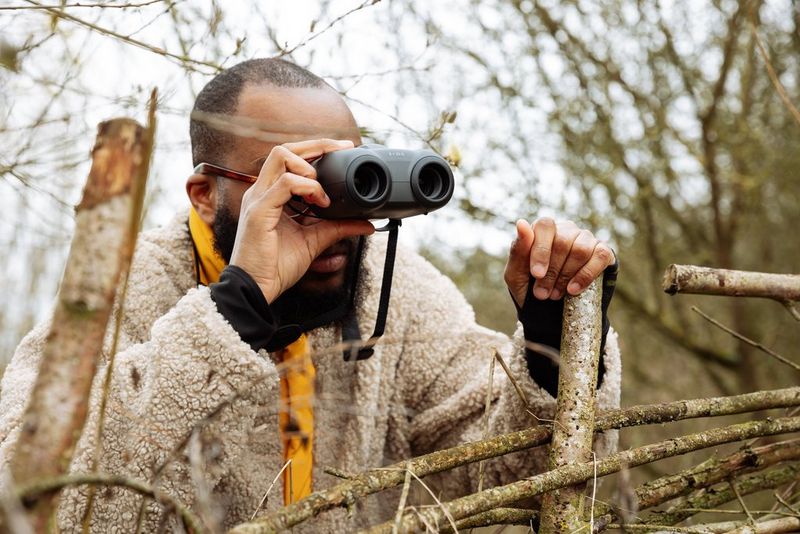 A man leans against a wooden fence holding a pair of Canon binoculars to his eyes.