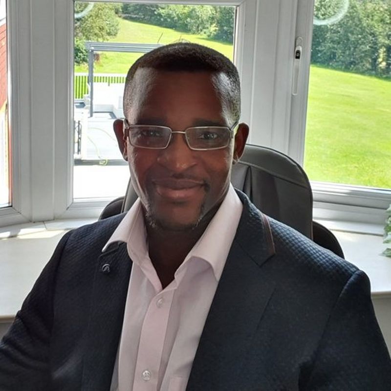A bespectacled man in a suit jacket and open necked white shirt sits in front of a window, through which you can see a lush green lawn.