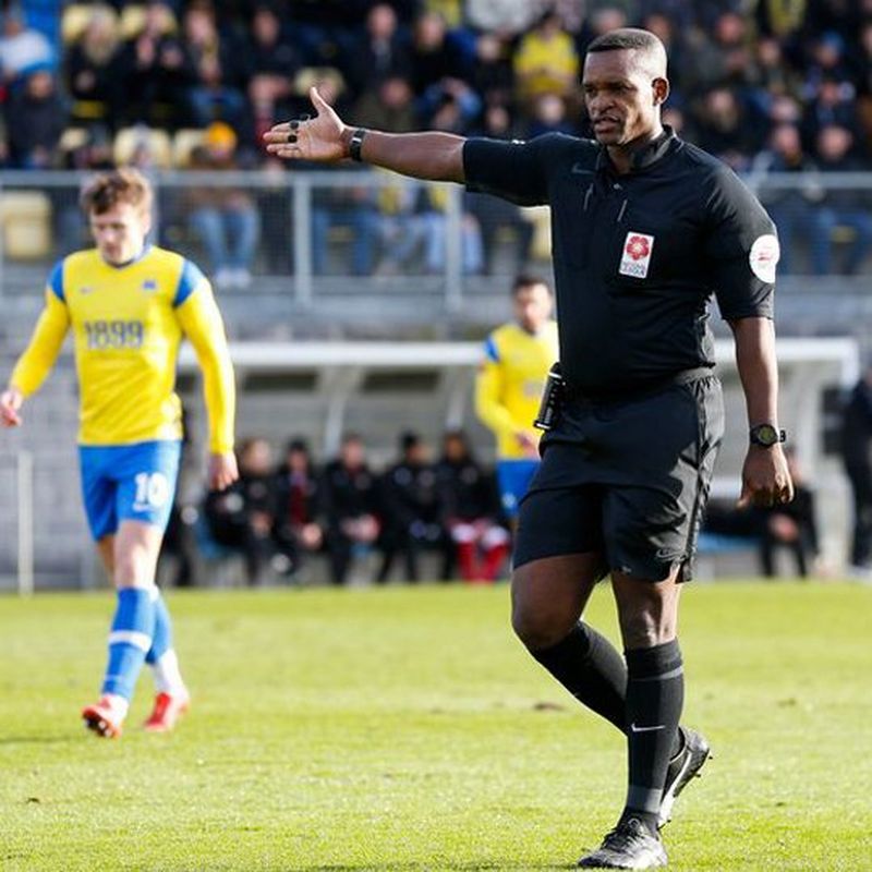 A man in a black referee’s uniform stands on a football pitch with his right arm outstretched. To his left and rear is a footballer wearing a yellow top, blue shorts and matching socks. There are blurred figures in the background who appear to be sat in the players dugout. Behind them are the crowd, who are watching the match.