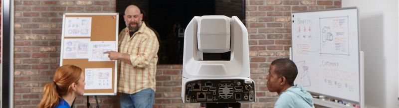 A man with a beard stands by a pin board, pointing at what is pinned on it. He wears a yellow checked shirt and blue jeans. In front of him, are the heads and shoulders of two people he is presenting to, who have their backs slightly turned to the camera. On the left is a woman with red hair and a bright blue blouse. On the right is a man with a pale blue hoodie. In the centre of the shot, at the head of the table, is the rear of a Canon PTZ camera.