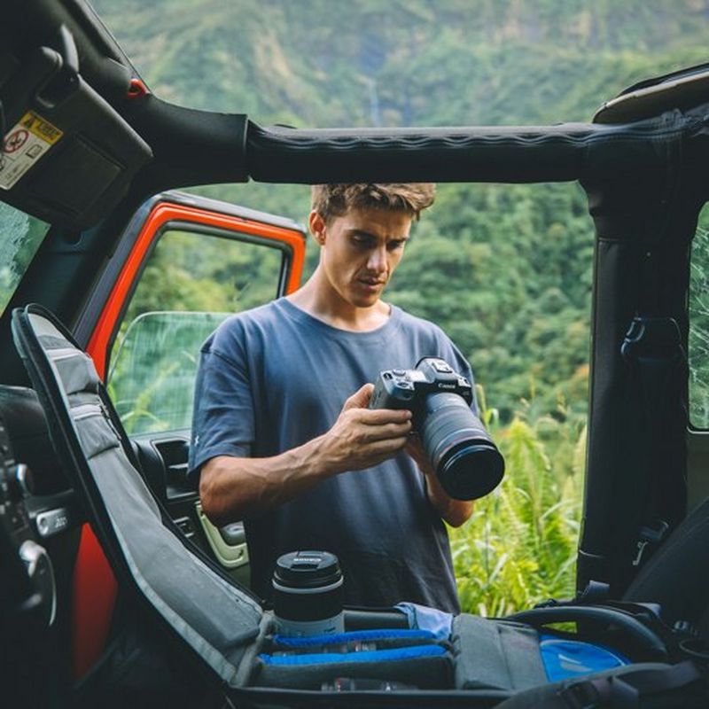 A man stands by the open door of an open-topped vehicle. In front of him is a case with a lens standing upright in it. He holds a camera, with a large lens attached and inspects the rear viewfinder.