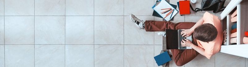 Taken from above, a teenage boy wearing a peach t-shirt and brown trousers sits on a tiled floor using a laptop. He has shelves behind him and stationery around him on the floor.