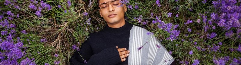 A bespectacled young man in a black and grey sweater lies in long grass, surrounded by bluebells. His right-hand rests on his chest at heart height. (© Boys by Girls Magazine)