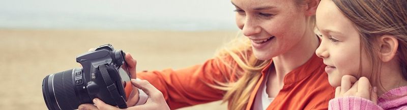 A woman in an orange top holds a Canon camera to show the rear display to her daughter, who wears a pink jumper. Although only their faces and shoulders can be seen, they appear to be against a background of a sandy beach.