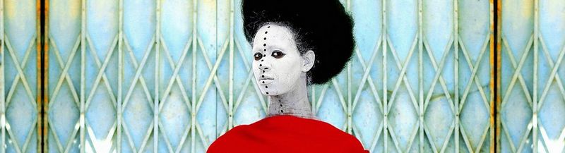 A woman stands in front of a rusting security grille. She wears a dramatic red cape and her head is framed by a halo of black hair. Her face is painted white and a line of black dots stretches from the top of her forehead to the base of her neck, which are inspired by traditional African body art.