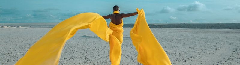 A woman walks on a sandy, flat surface towards the ocean. Her arms are stretched wide, and she wears bright yellow chiffon that trails behind her, blowing in the wind and creating a strong contrast to the blue sky.