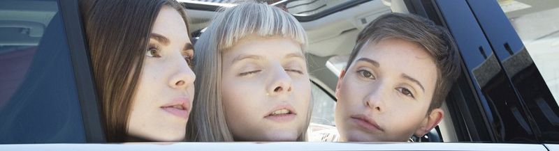 A close-up of three women leaning their heads out of a car window. The woman on the left has long brown hair, the woman on the right has short brown hair and, in the middle, the woman is blonde and has her eyes closed and head raised gently skywards.