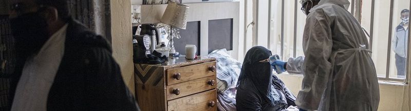 A man dressed in white PPE takes the temperature of a women as she sits up in bed. She is wearing a black niqab and on one side of her is a high brown wood chest of draws with six drawers and a lamp and assorted objects on top. On the other side is a barred window, through whIch you can see the figure of a man wearing a face covering. On the far left of the photo is the side view of a man wearing a white kameez and white topi. He also wears a black protective face covering.