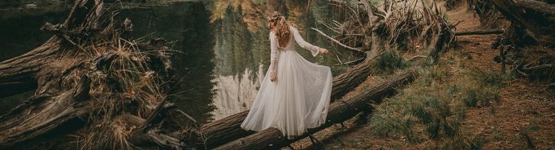 A women with long hair in her wedding dress is balancing on fallen trees on the edge of a give in a forest.