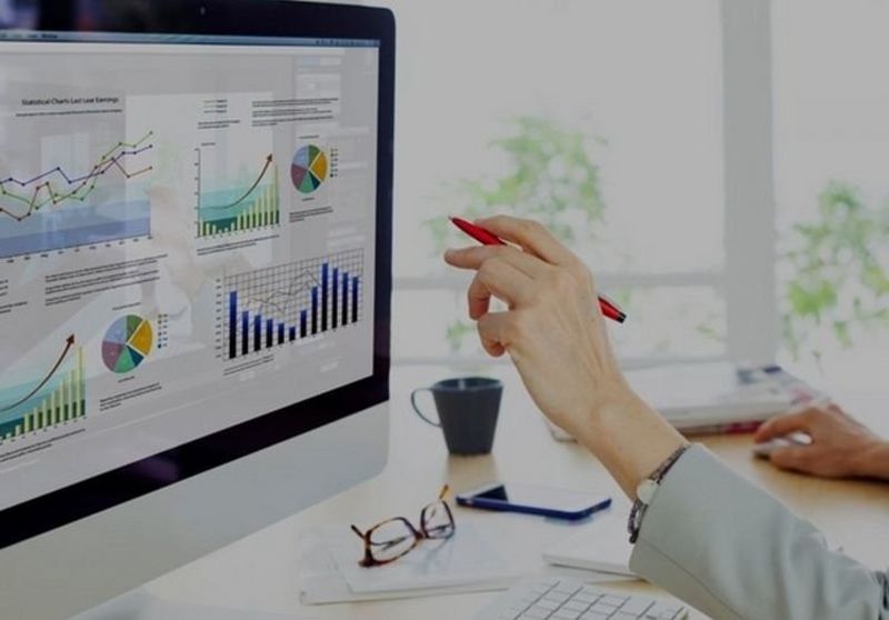Close-up of a woman’s hand holding a red pen and pointing to a large computer screen made up of pie charts and graphs.