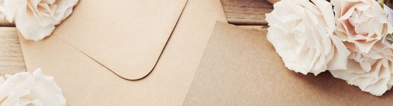 A brown paper envelope and the edge of a brown kraft paper card, on a table, surrounded by white rose-like flowers.