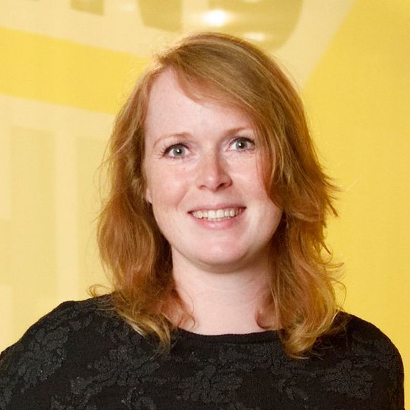 A smiling, red haired woman in a black top stands in front of a bright yellow background.