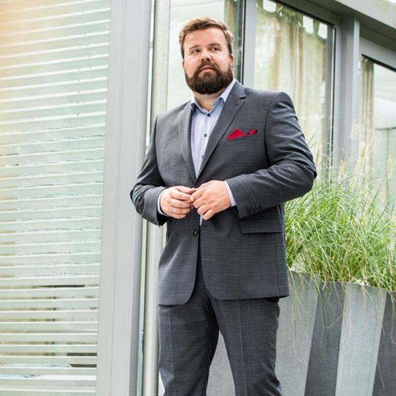 A bearded man stands in front of a windowed building, wearing a grey suit, blue shirt and has a red pocket square. There is a green plant to his right.