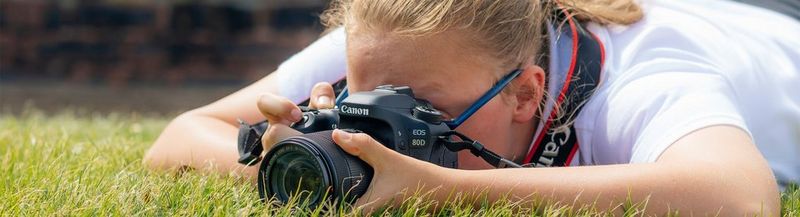 A girl lies on the grass, her face obscured by a Canon EOS 80D, as she takes a shot