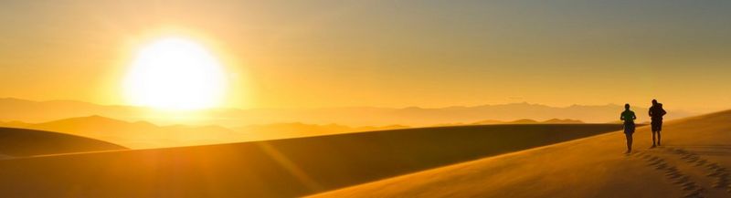 On the right, a small silhouette of a couple walking across golden sand dunes at dusk, leaving footprints behind them. Far on the horizon, a huge bright sun begins to set.
