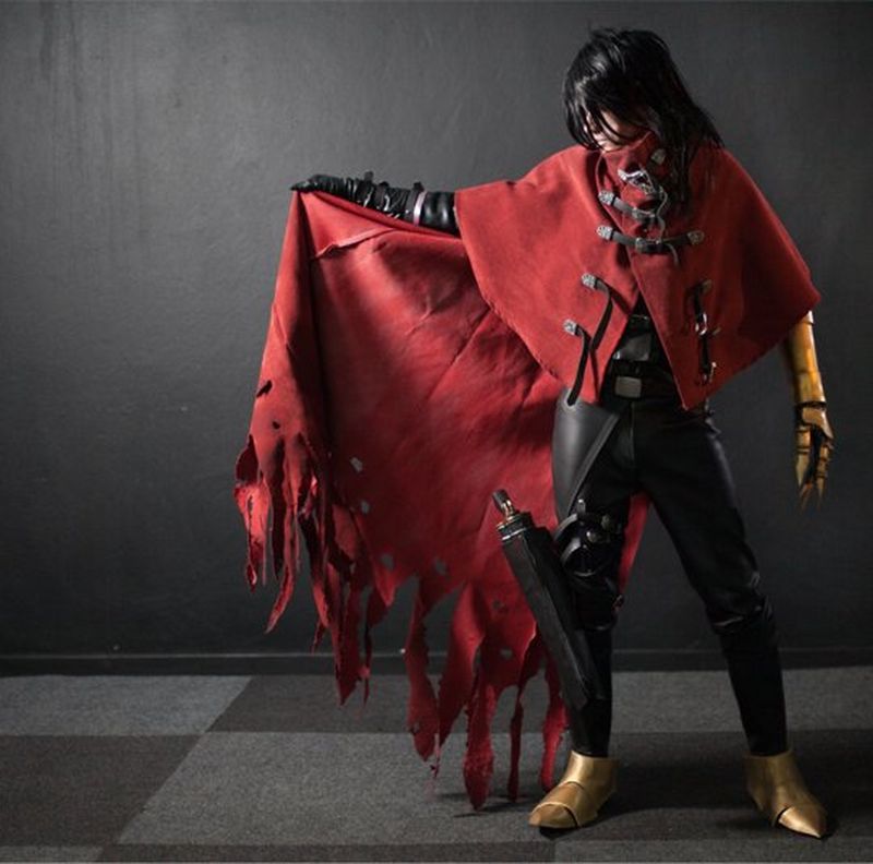 A cosplayer stands against a dark background, face covered, lifting a red cape.