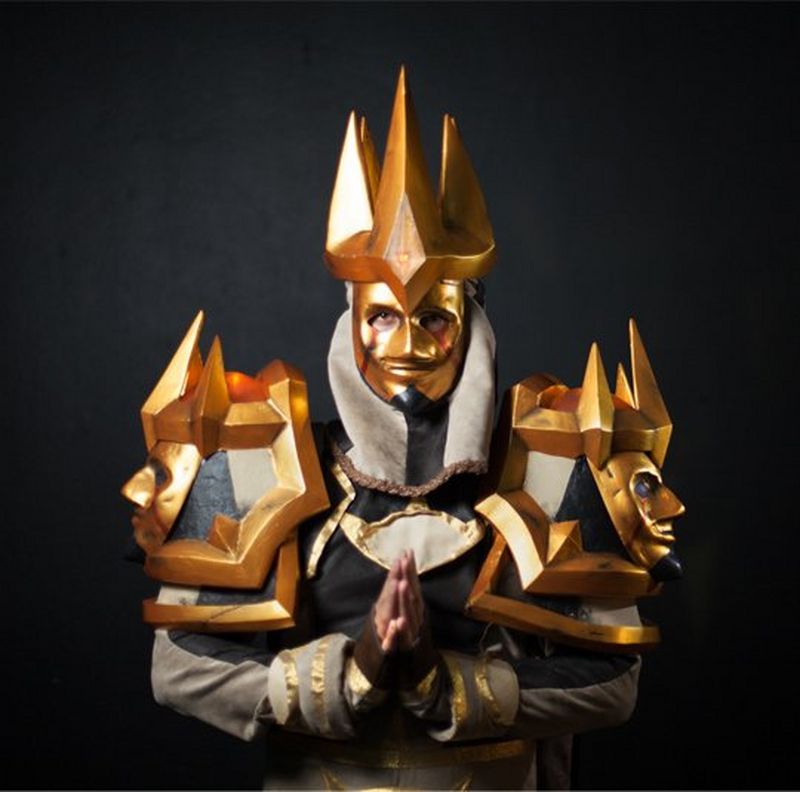 An elaborate costume of gold and silver, with gold masks as epaulets. The cosplayer stands with hands on prayer position.