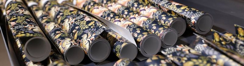 Eight rolls of wallpaper (and more just out of shot), photographed side by side, from the end of the rolls. They are patterned with leaves and flowers on a black background.
