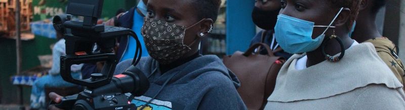 Two women – Jane Wanjiru Mumbi and Mercy Owegi. Both are wearing face coverings and only with head and shoulders visible, are facing left as though looking at something. The woman on the furthest left holds a Canon video camera.