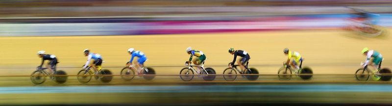 A photograph of seven professional cyclists in a line along what must be a velodrome. However, the photograph has substantial motion blur and all that can be seen of the background are streaks of yellow, blue and pink.
